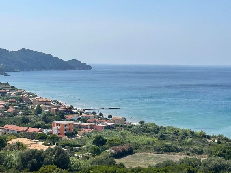 Panorama-Ansicht der Küste von Arillas mit blauem Meer, bewaldeten Hängen und verstreuten Häusern unter klarem Himmel.