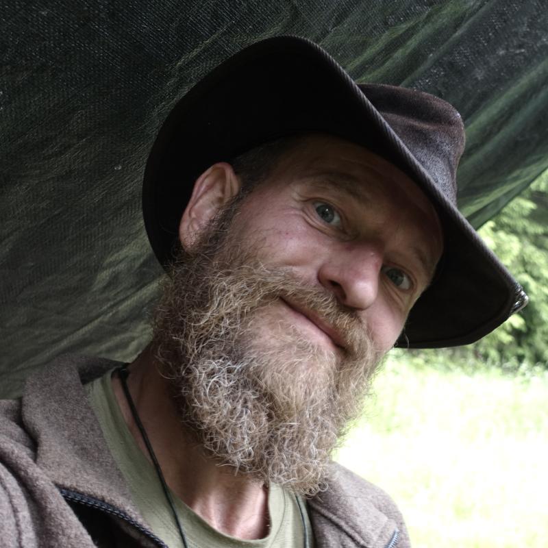 Nahaufnahme eines bärtigen Mannes mit dunklem Hut, der unter einer Plane (Tarp) hervor lächelnd und aufmerksam in die Kamera blickt.