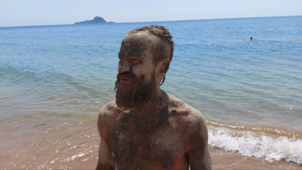 Florian Köhler, Gesicht und Oberkörper vollständig mit Schlamm bedeckt sind, steht am Meeresufer.