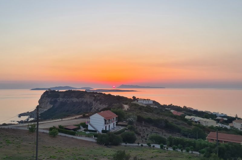 Blick von einer Anhöhe auf ein weißes Haus mit Ziegeldach an der Küste von Korfu während eines leuchtend roten Sonnenuntergangs über dem Meer und fernen Inseln.