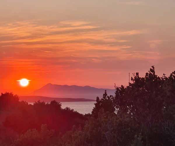 Ein intensiver, tiefroter Sonnenuntergang über dem Meer auf Korfu, im Vordergrund die Silhouette mediterraner Vegetation.