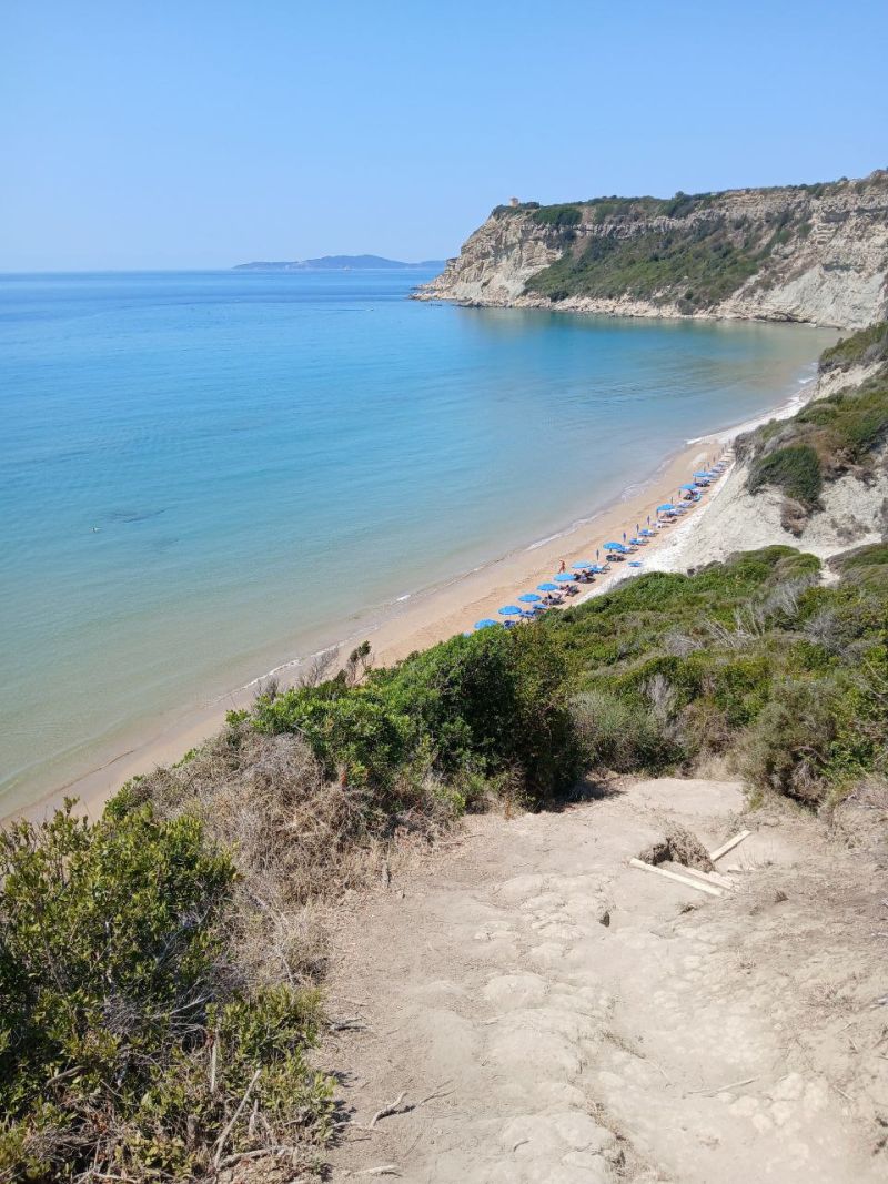 Ein sandiger Pfad führt von einer Anhöhe hinunter zu einem hellen Sandstrand mit blauem Wasser und markanten Felsklippen unter klarem Himmel.