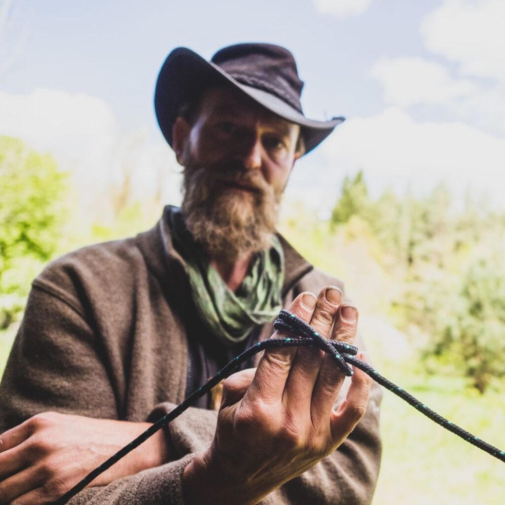 Ein Mann mit Vollbart, Hut und Wolljacke hält eine dunkle Kordel in der Hand und demonstriert vor grünem Hintergrund Knotentechniken im Rahmen der Wildnispädagogik.