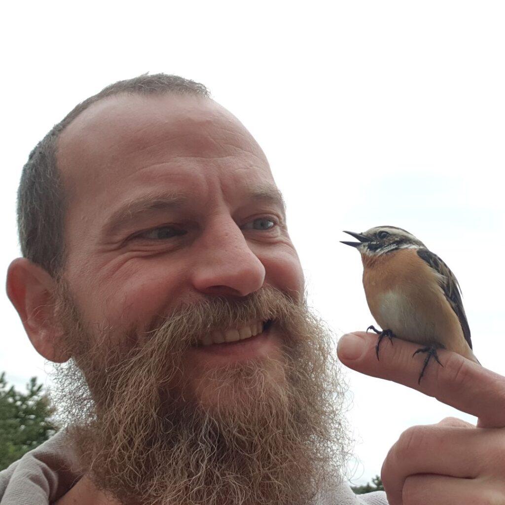 Ein bärtiger Mann lächelt freundlich, während ein kleiner, braun-schweifiger Vogel vertraut auf seinem Zeigefinger sitzt und zwitschert.
