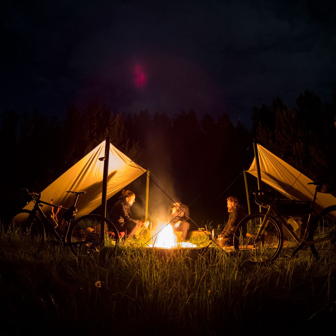 Drei Personen sitzen nachts bei Dunkelheit gemütlich an einem hell lodernden Lagerfeuer, umgeben von zwei aufgespannten Tarps und geparkten Fahrrädern im Gras.