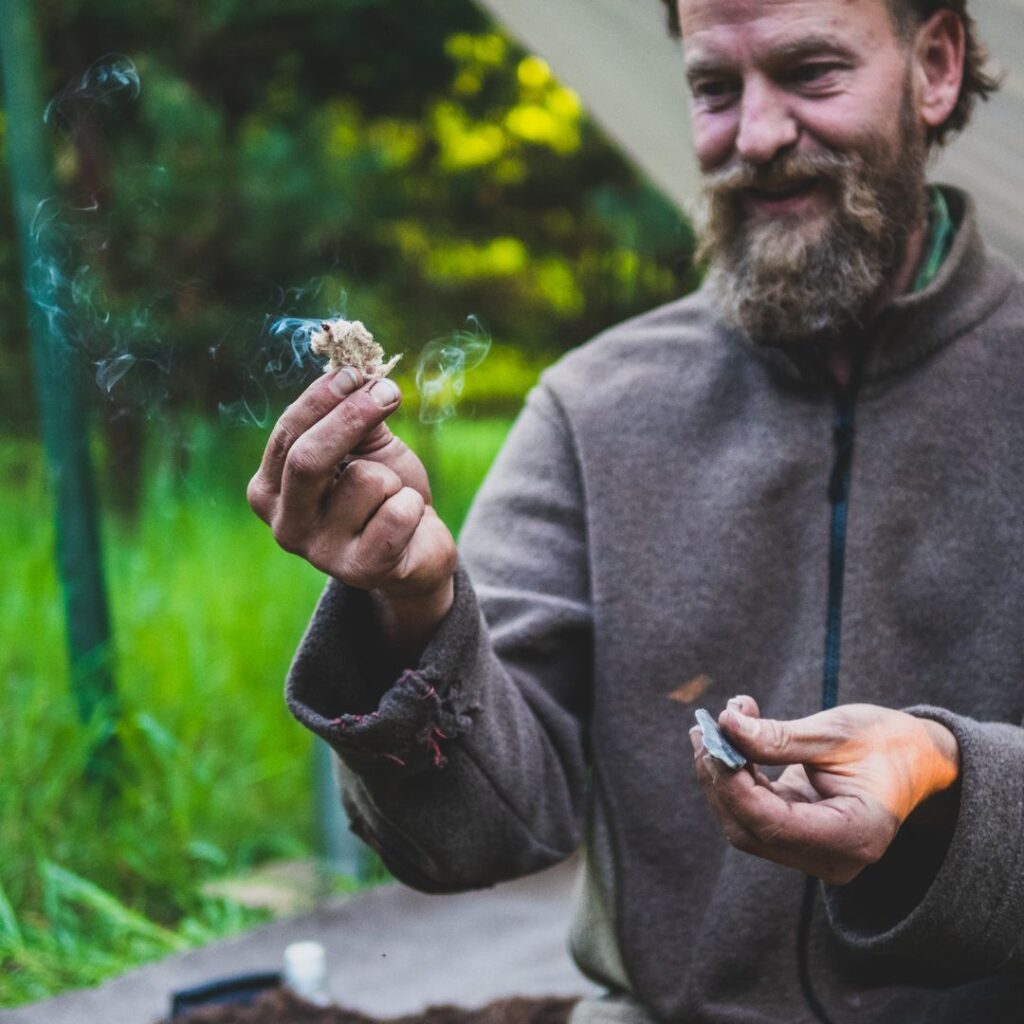 Nahaufnahme eines Mannes mit Vollbart und brauner Wolljacke, der ein rauchendes, glimmendes Stück Zunder in der Hand hält und beim ursprünglichen Feuermachen im Wald lächelt.