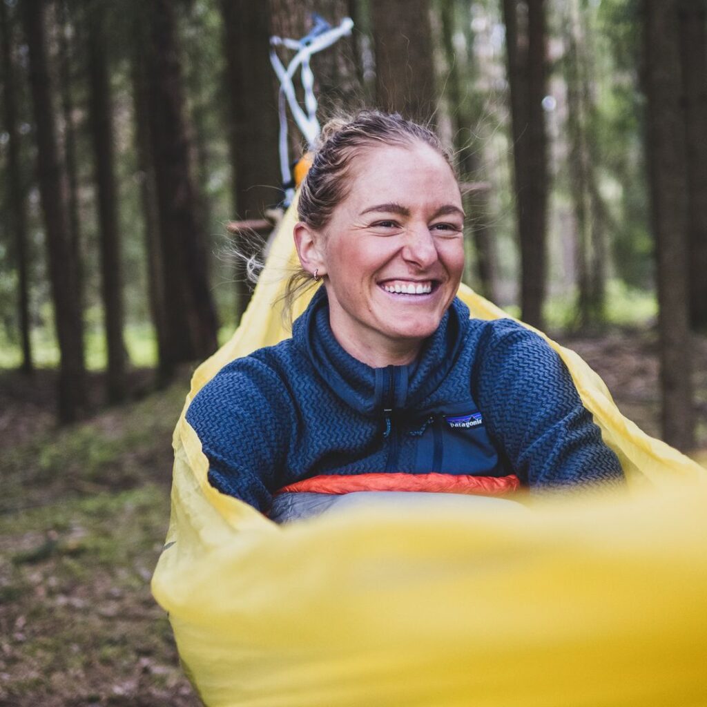 Eine glückliche Frau lacht strahlend, während sie gemütlich in einer gelben Hängematte zwischen dichten Nadelbäumen im Wald liegt.