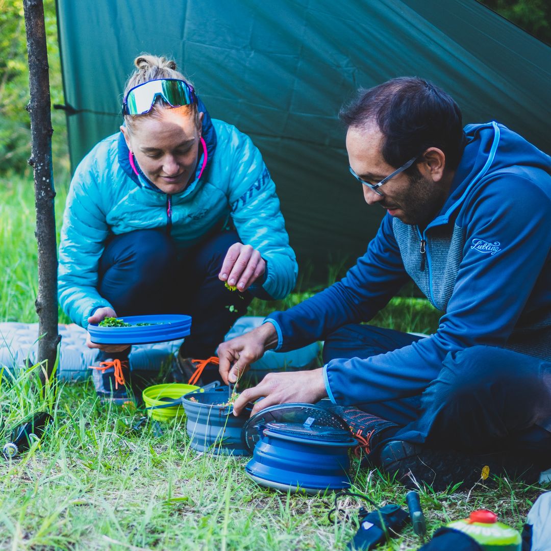 Eine Frau in blauer Daunenjacke mit Stirnband und ein Mann in blauer Fleecejacke bereiten gemeinsam unter einem grünen Tarp im Gras eine Mahlzeit mit Outdoor-Kochgeschirr zu.