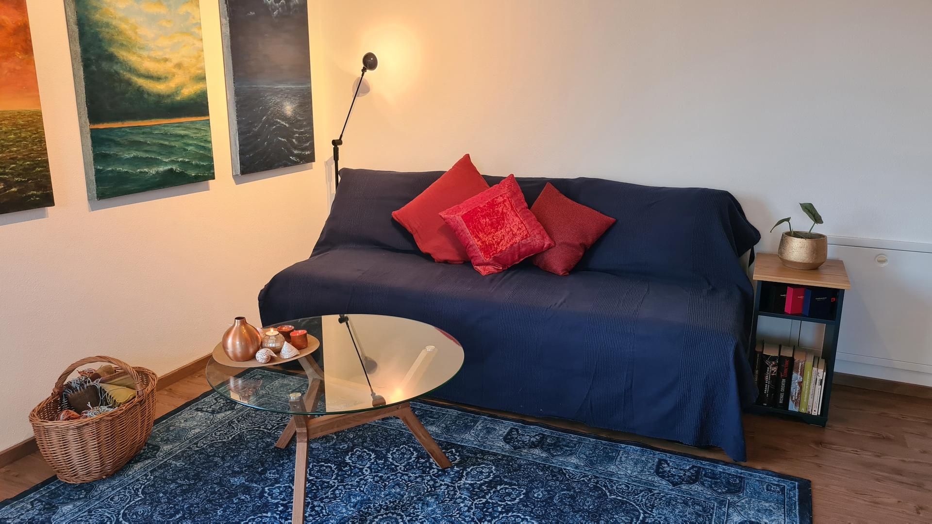 Sofa mit blauer Tagesdecke und roten Dekokissen - Gemälde an der Wand und Deko auf Glastisch