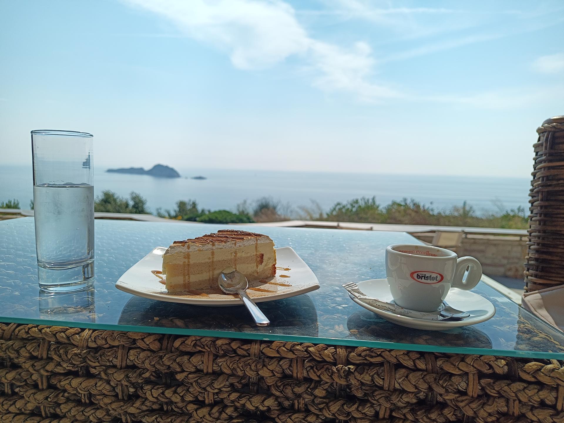 Ein Tisch mit Kuchen, Espresso und Wasser vor einem weiten Blick auf das offene Meer.