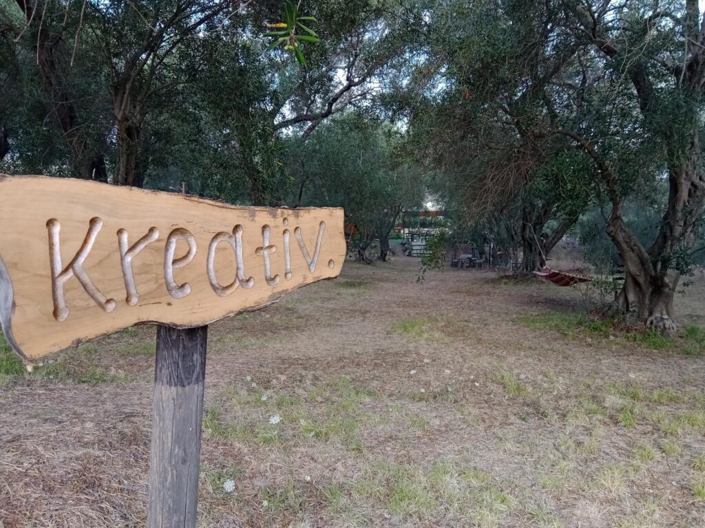 Ein Olivenholz-Schild mit Aufschrift „Kreativ“ weist in den Olivenhain mit Hängematten des Ouranos Clubs auf Korfu