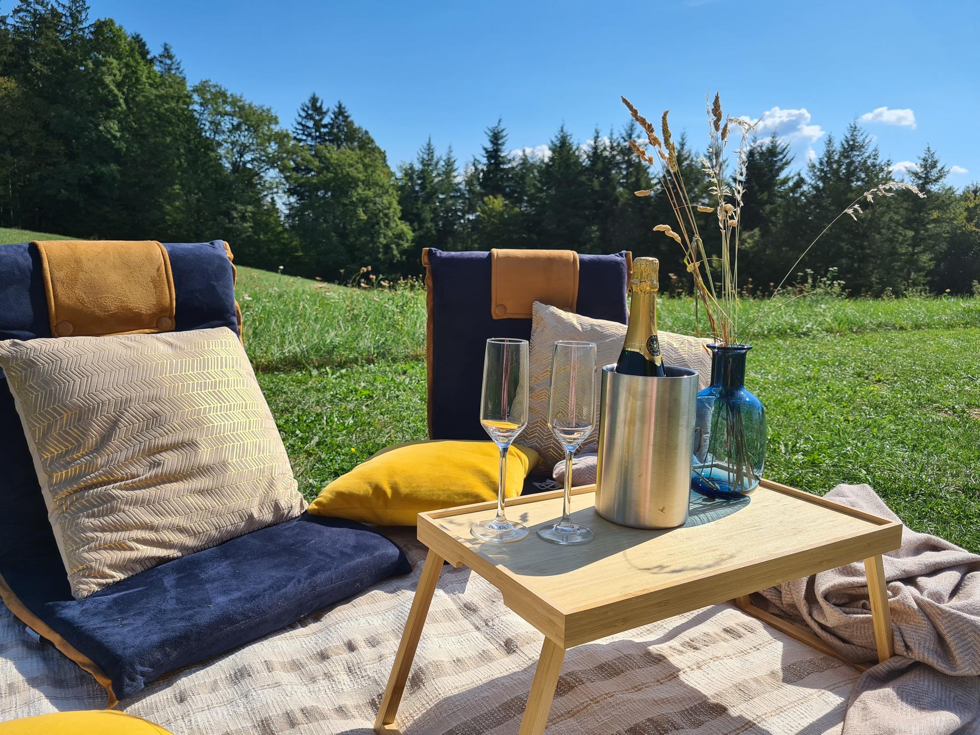 Ein Stelltischchen auf einer Picknickdecke vor einem Waldrand mit Sekt und Gläsern, daneben zwei gepolsterte Bodenstühle und Kissen