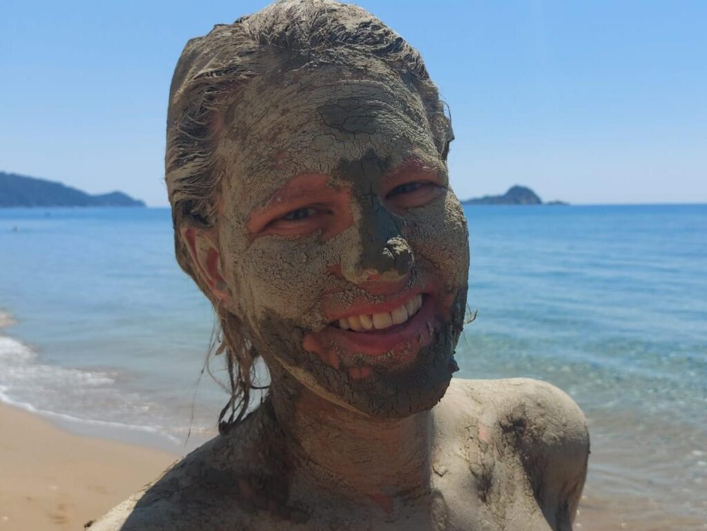Eine Frau am Strand auf Korfu lächelt glücklich in der Sonne - mit Heilschlamm im Gesicht